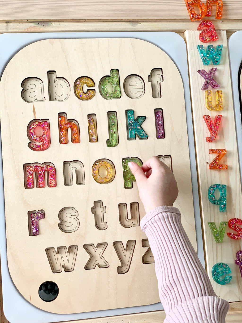 Child's hand placing colorful resin lowercase letters into wooden alphabet puzzle board insert on table