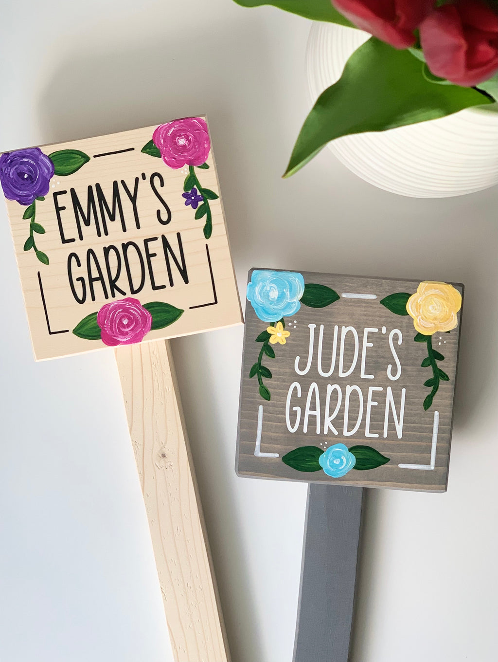 Two small square wooden garden signs on stakes painted with names and colorful floral decorations on wood backgrounds