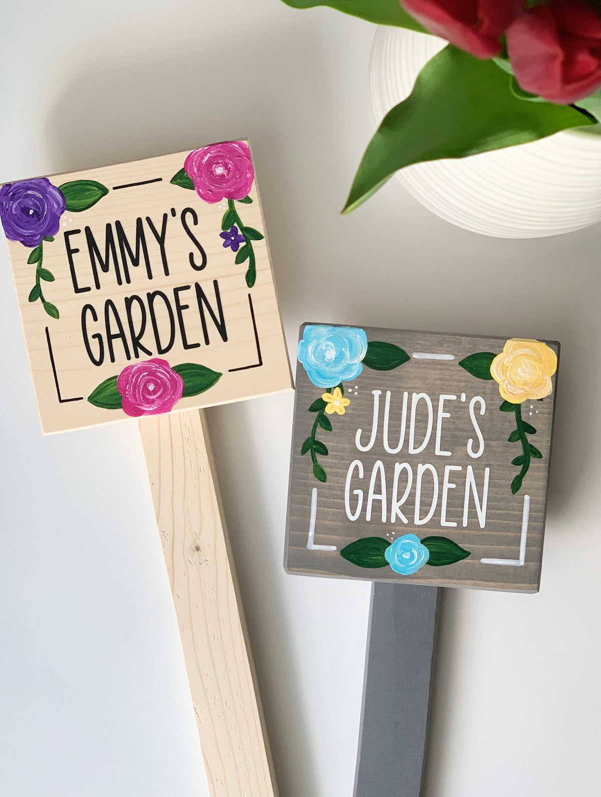 Two small square wooden garden signs on stakes painted with names and colorful floral decorations on wood backgrounds