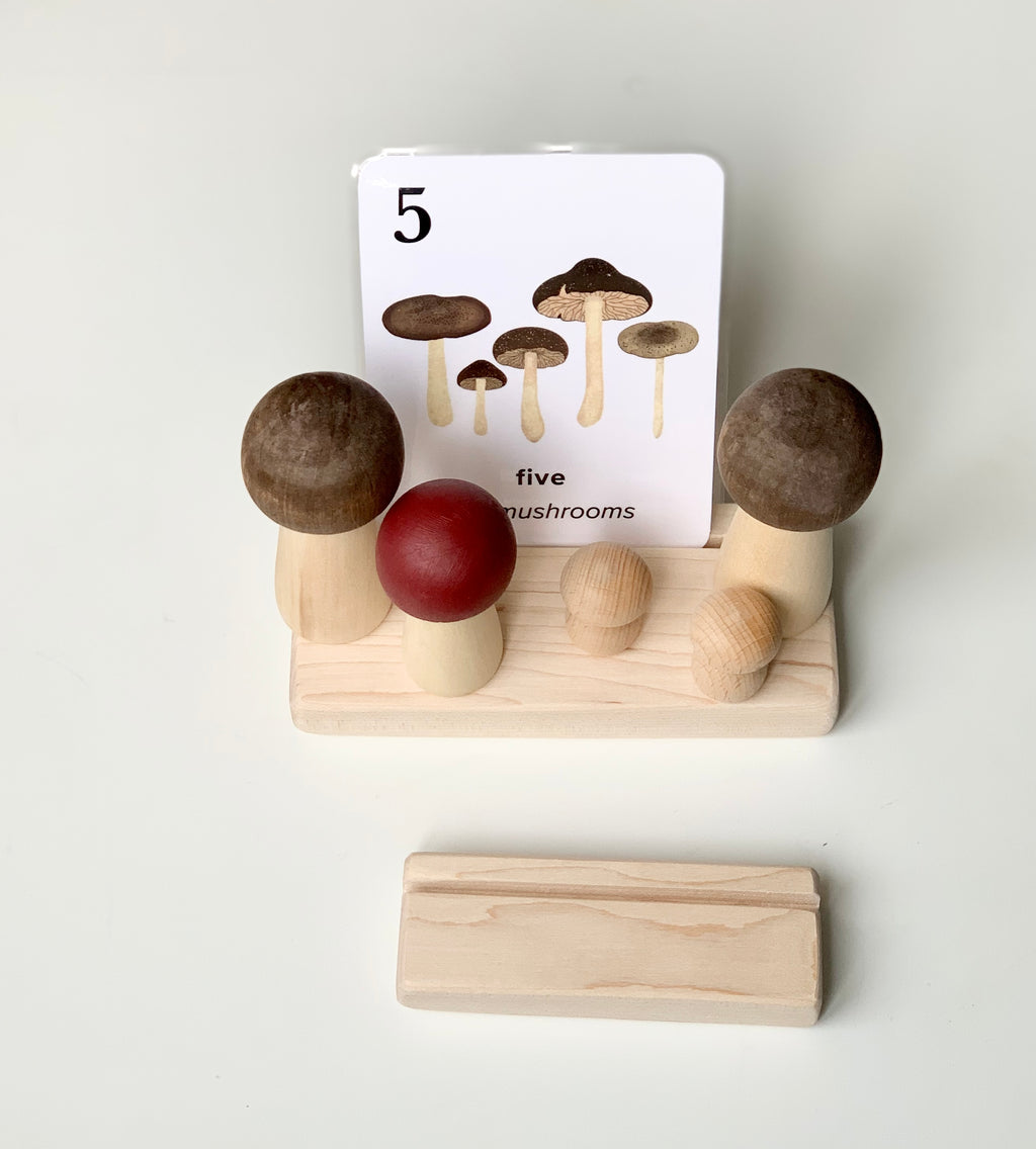 Natural wooden flash card holder with wide slots holding mushroom-themed flash card and wooden mushroom figures on white surface
