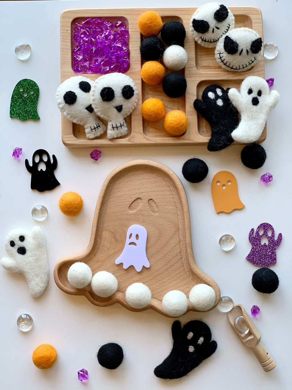Wooden ghost-shaped sensory tray with felt skulls, ghosts, orange and black balls, and purple decorative gems on white surface