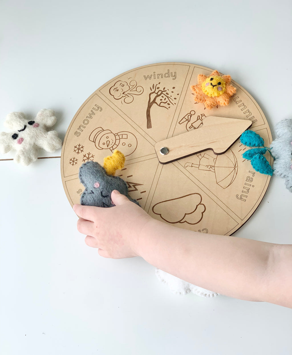 Round wooden weather wheel with engraved weather icons and child’s hand holding felt thunderstorm figure on white background