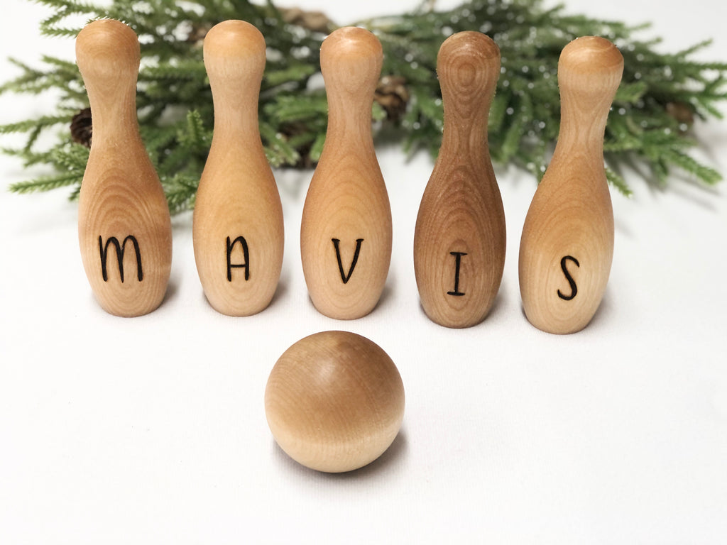Wooden personalized toddler bowling set with five skittles spelling MAVIS and a wooden bowling ball on a white surface