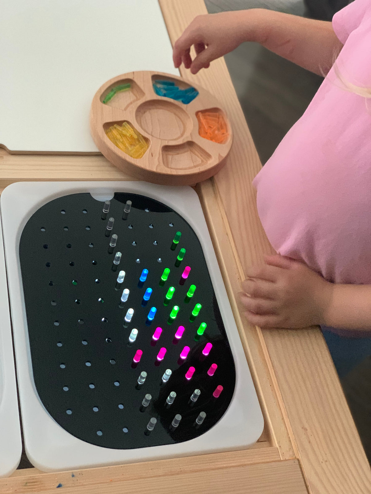 Black geometric peg board with colorful light pegs inserted on wooden Flisat table with child’s hands nearby