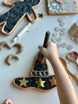 Child's hand holding brush over wooden witch hat plate filled with black rice and decorated with gold stars and clear stones on white surface