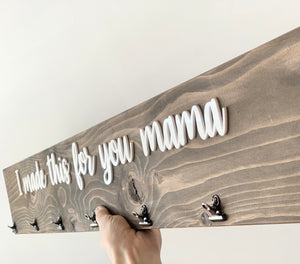 Rustic wooden sign with white cursive letters saying I made this for you mama and black clips for displaying artwork