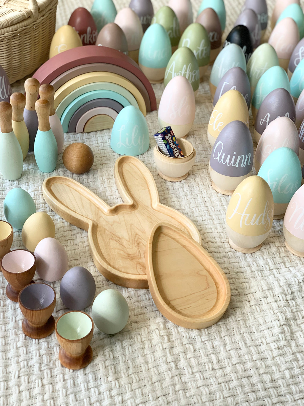 Wooden fillable Easter eggs in pastel colors with cursive names, wooden bunny tray, egg cups, and colorful wooden rainbow toy on fabric surface