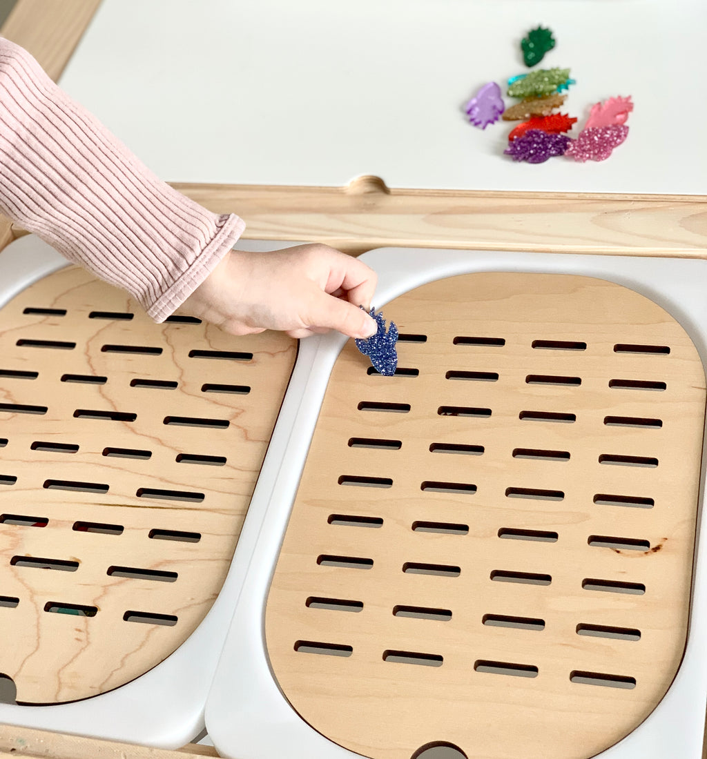 Hand placing blue glittery shape into Flisat table top insert slot with colorful play pieces on light wood surface