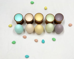 Five natural wood eggs in pastel colors with matching wooden cups and scattered small colored eggs on white background