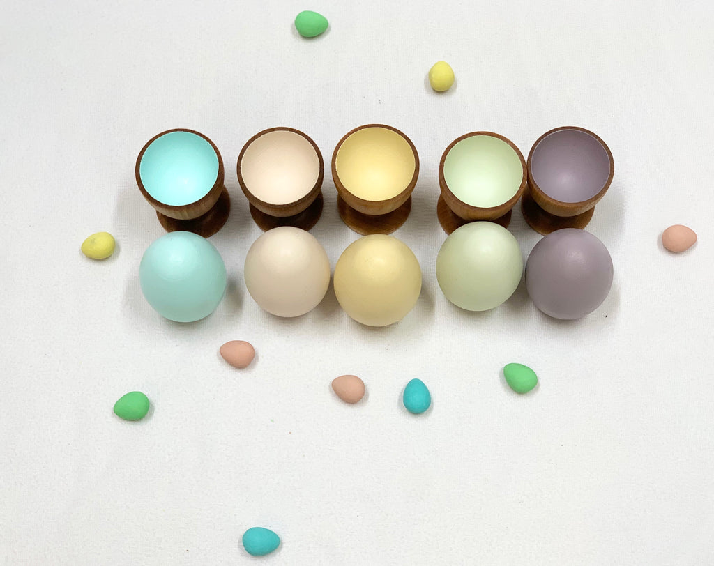 Five natural wood eggs in pastel colors with matching wooden cups and scattered small colored eggs on white background