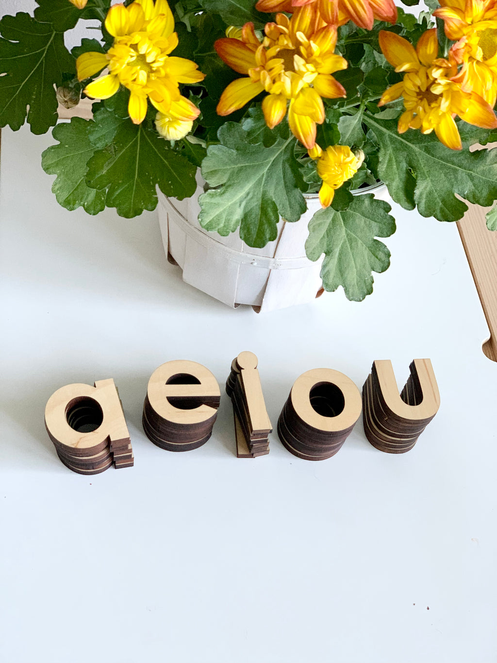 Natural wooden lowercase alphabet letters stacked to spell vowels on white surface next to yellow and orange flowers