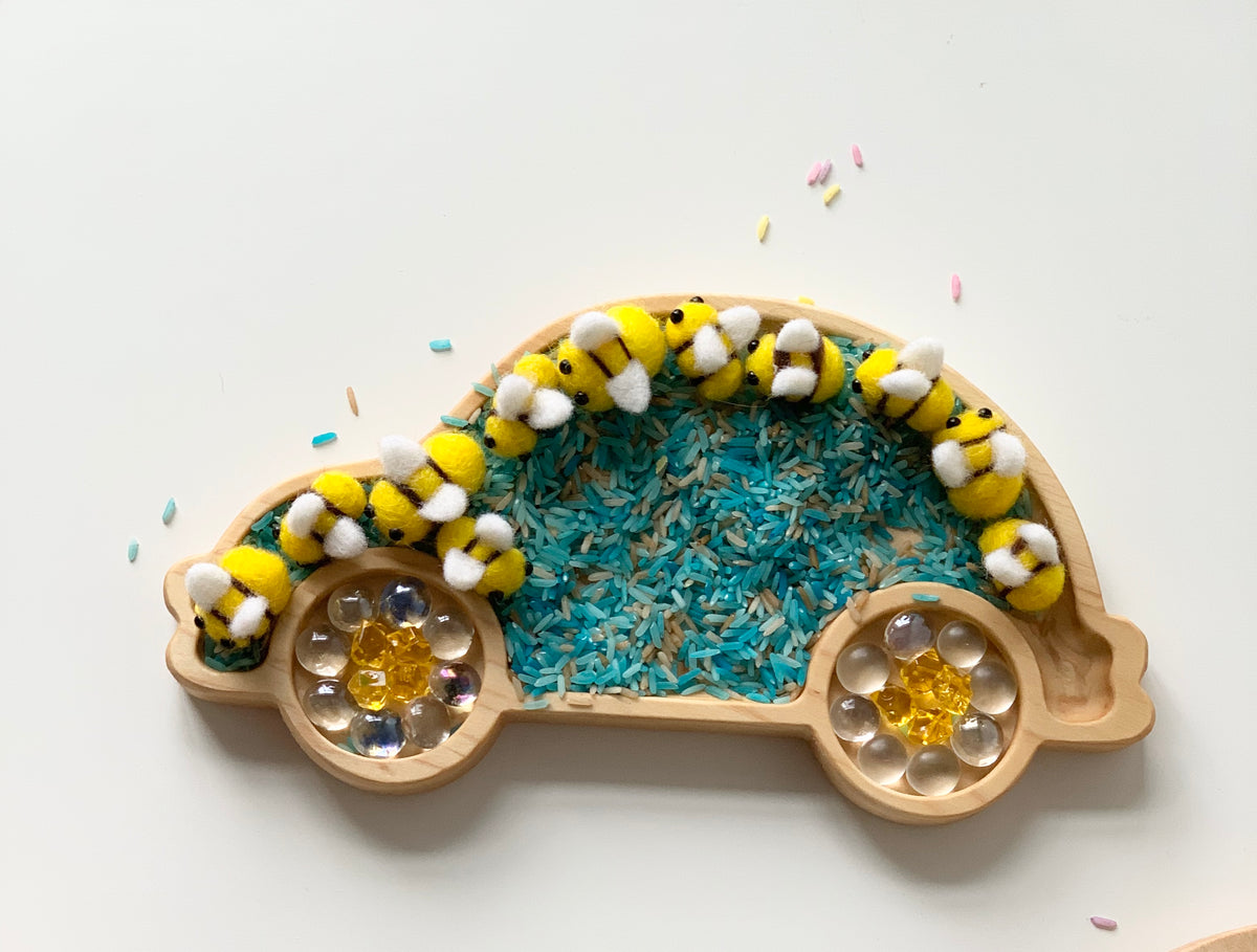 Natural wooden car-shaped sensory tray with colorful rice, glass beads, and a yellow caterpillar toy on white background