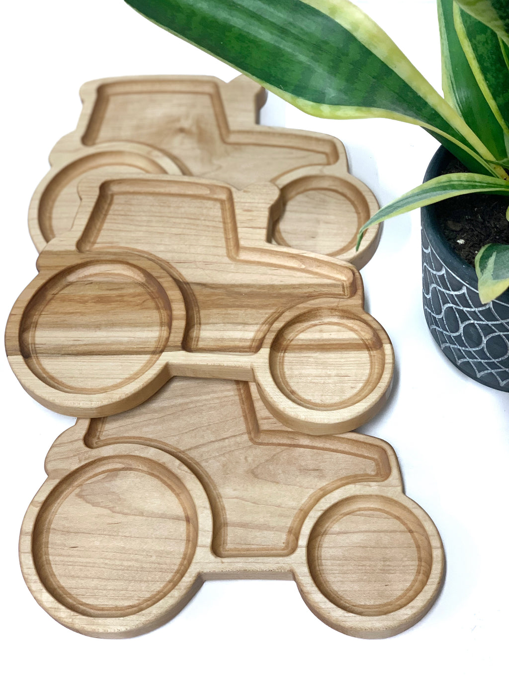 Three natural wooden tractor-shaped sensory trays stacked near green potted plant on white surface