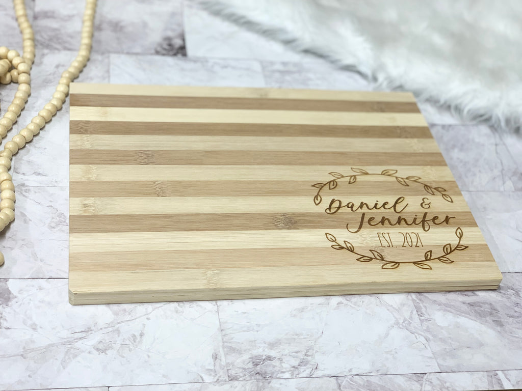 Rectangular striped wooden cutting board with personalized engraved names and date inside wreath design on bottom right corner