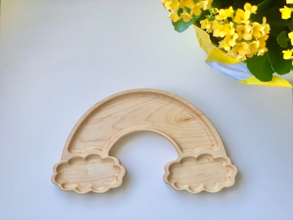 Natural wooden sensory tray shaped like a rainbow with cloud compartments on light background beside yellow flowers