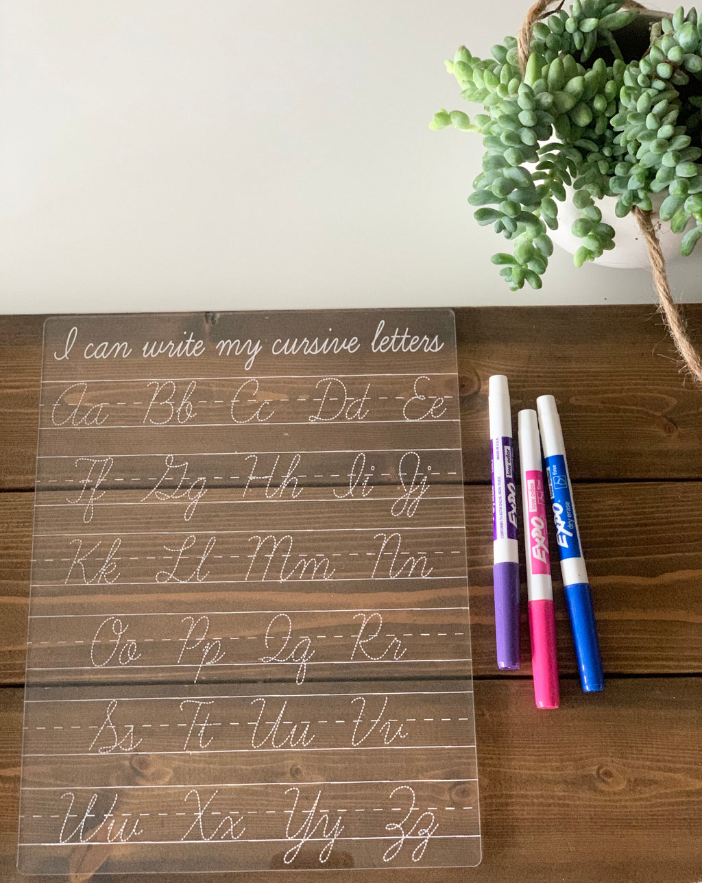 Transparent acrylic dry erase board engraved with cursive alphabet letters and three colored markers on wooden surface