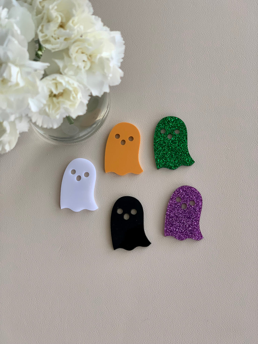 Set of five Halloween ghost laser cut acrylic shapes in white, orange, black, green glitter, and purple glitter on neutral surface