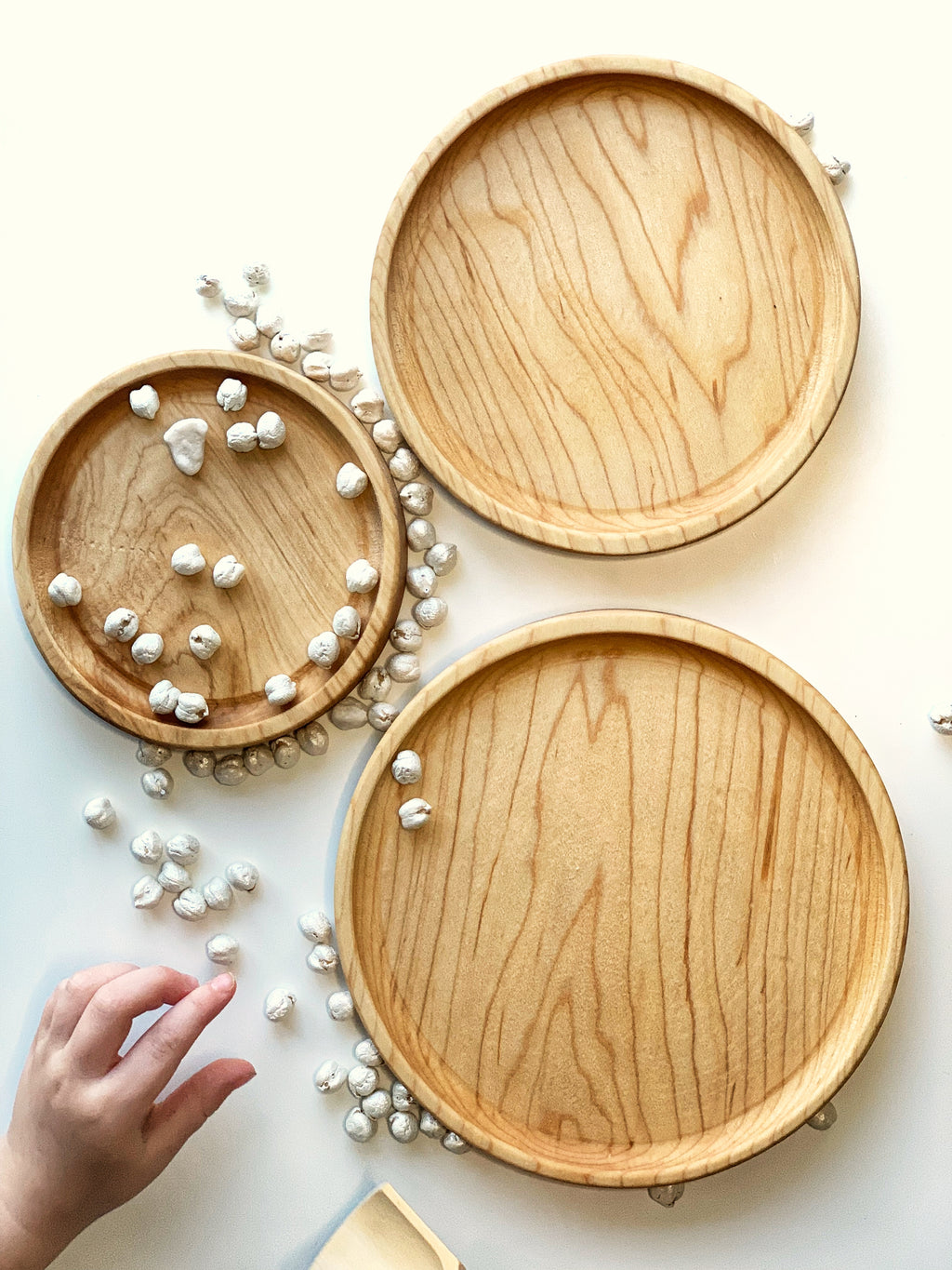 Three natural wooden nesting round plates with scattered small white balls on white surface and child hand reaching in frame