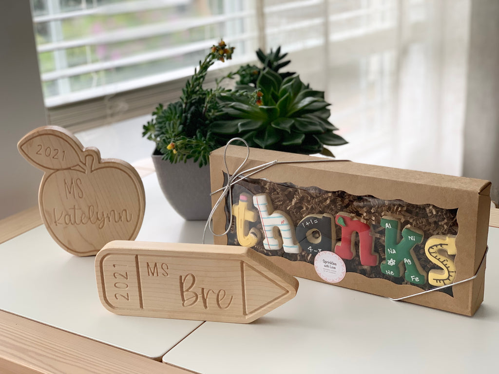 Wooden personalized teacher name plates shaped like an apple and pencil next to colorful block letters spelling THANKS in gift box