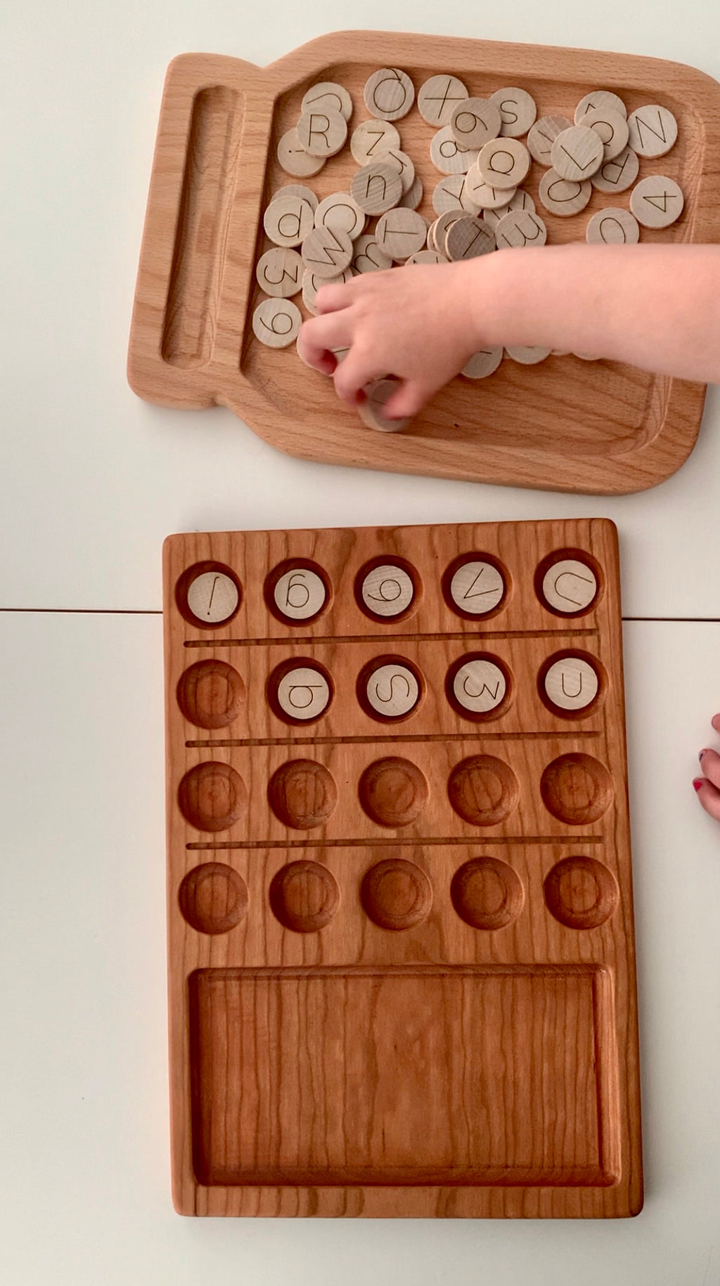 Natural wooden 20 frame sensory tray with engraved alphabet discs scattered on matching wooden tray held by child’s hand on white surface