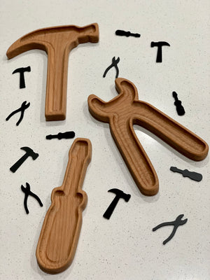 Wooden tool-shaped trays with black acrylic loose parts in hammer, wrench, screwdriver, and plier shapes on white surface