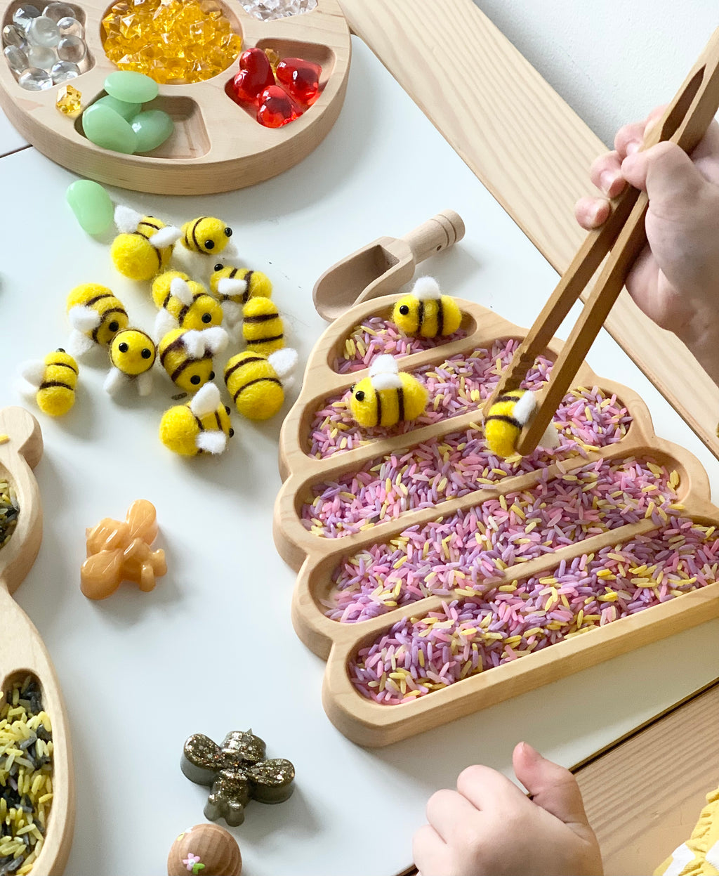 Bee hive shaped wooden sensory tray filled with colorful rice and small felt bee toys with child using wooden tongs