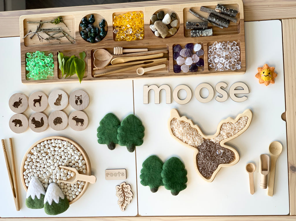Wooden sensory tray with moose head shape, natural objects, wooden letters, felt trees, and small wooden tools on white surface