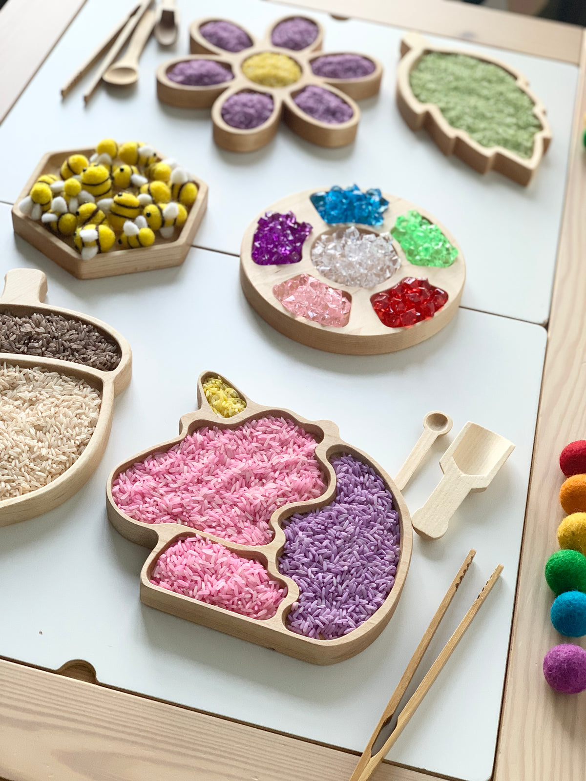 Wooden sensory trays shaped like a unicorn, leaf, flower, acorn, and hexagon filled with colorful sensory materials