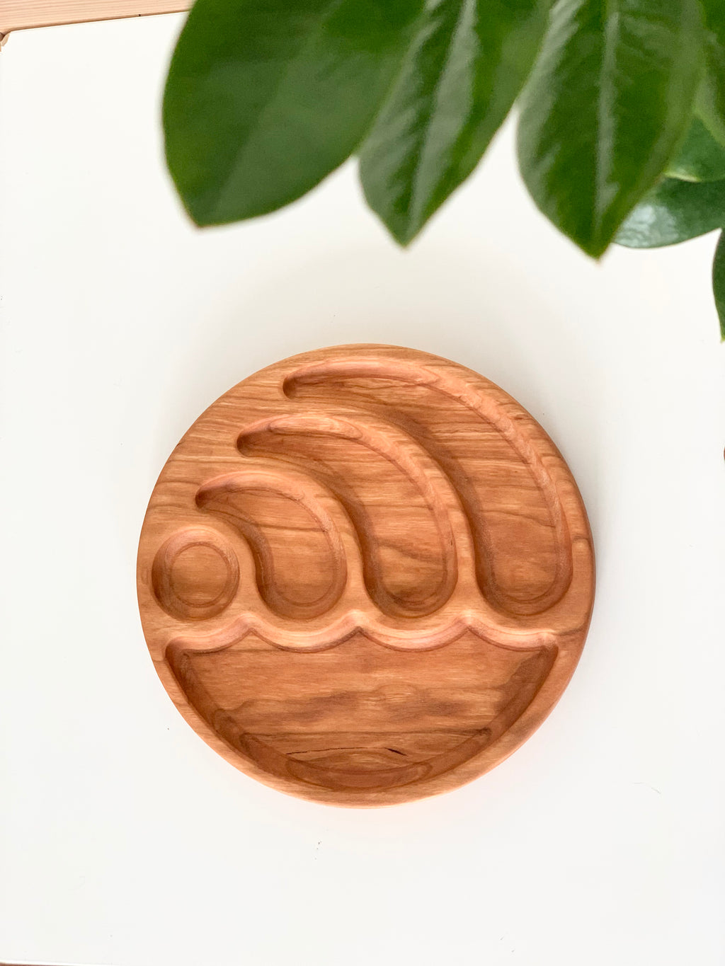 Round wooden sensory tray carved with wave shapes on white surface with green leaves overhead