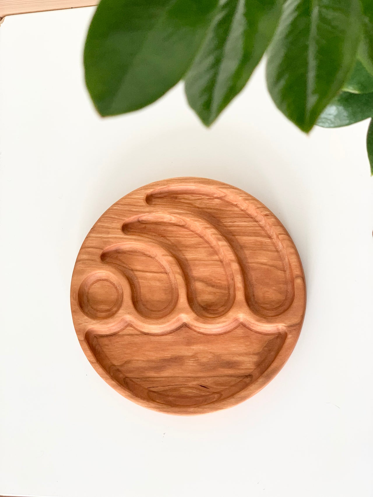Round wooden sensory tray carved with wave shapes on white surface with green leaves overhead