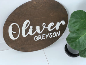 3D round wooden name sign with white cursive and block letters on dark stained wood background near green plant