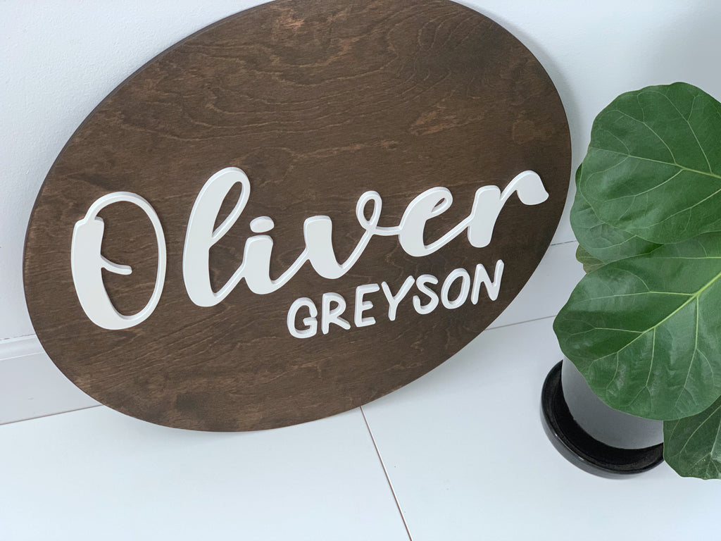 3D round wooden name sign with white cursive and block letters on dark stained wood background near green plant