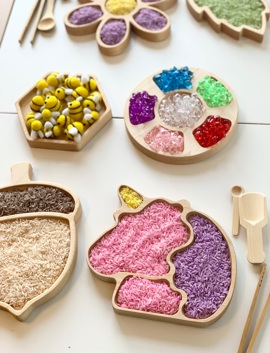 Natural wooden sensory tray shaped like a unicorn filled with colorful rice and surrounded by other wooden trays and tools