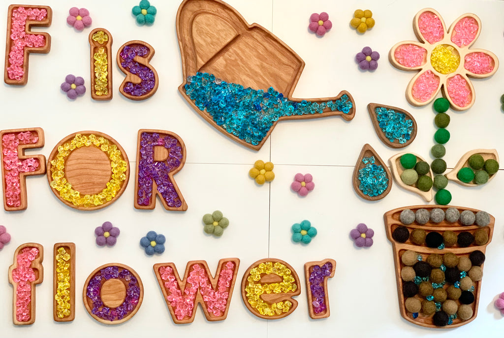 Wooden watering can sensory tray filled with blue beads surrounded by a flower tray and colorful felt flowers on white surface