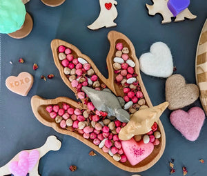 Wooden dinosaur footprint plate filled with pink and beige sensory beads on dark surface with heart and dinosaur toys