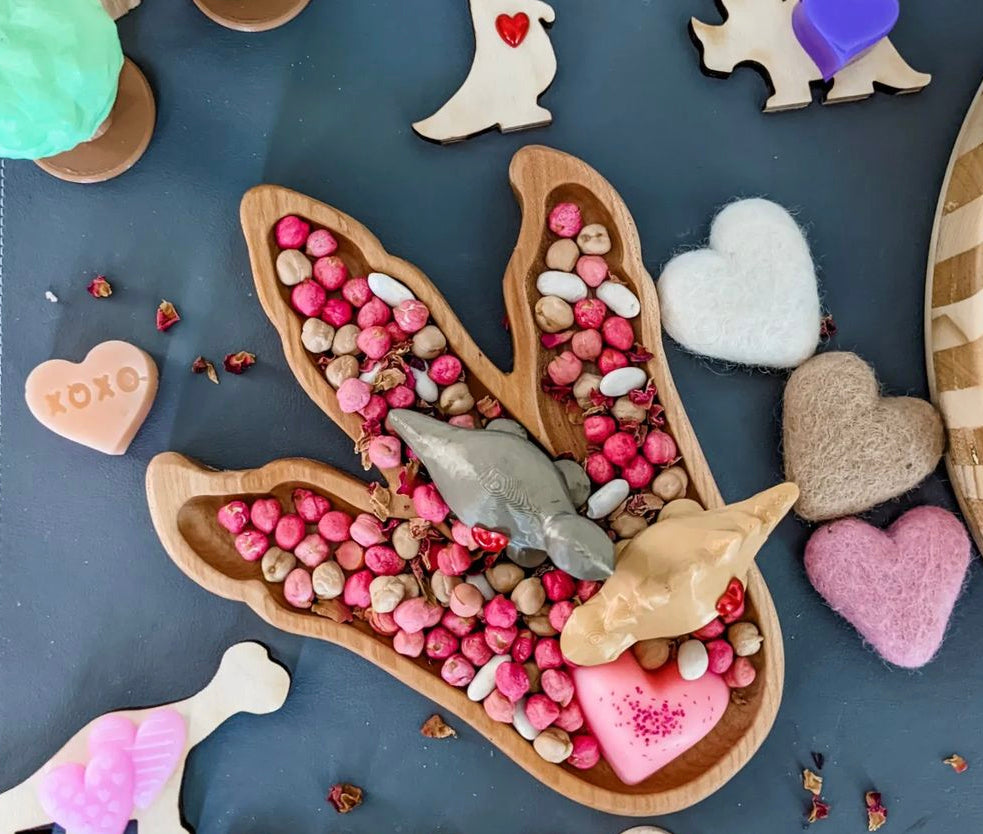Wooden dinosaur footprint plate filled with pink and beige sensory beads on dark surface with heart and dinosaur toys