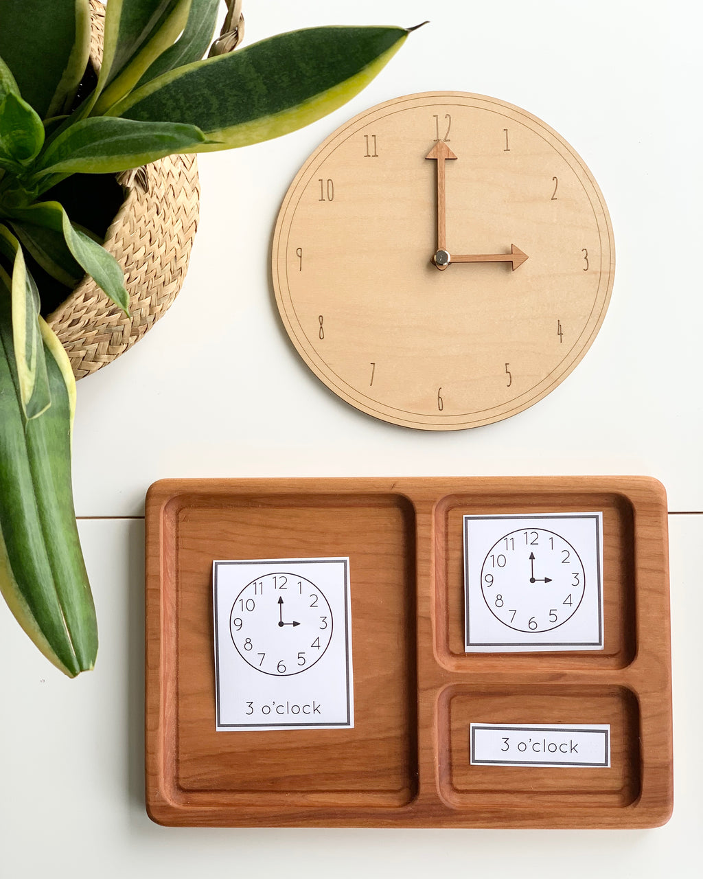 Natural wooden play learning clock with movable hands and matching time cards on wooden tray near green plant