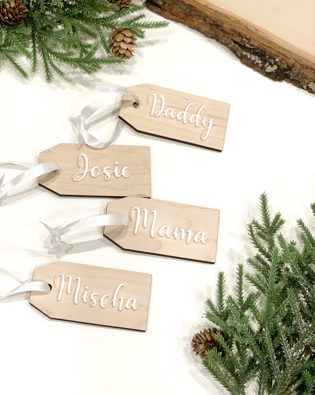Natural wooden 2x4 inch Christmas stocking and gift tags with white cursive names and ribbon on white background with pine branches