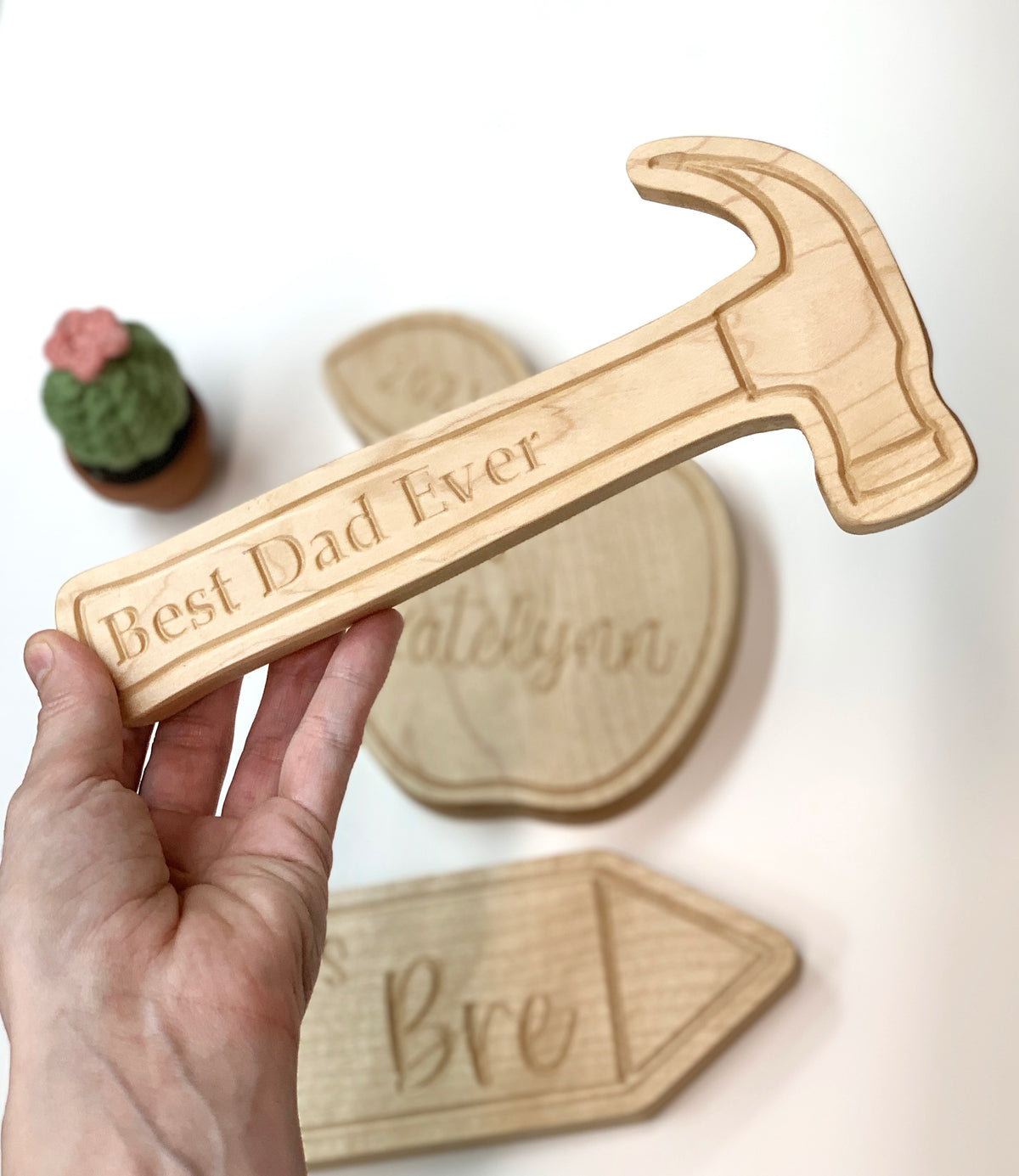Light natural wooden hammer-shaped plaque engraved with "Best Dad Ever" held above engraved wooden name signs on white background