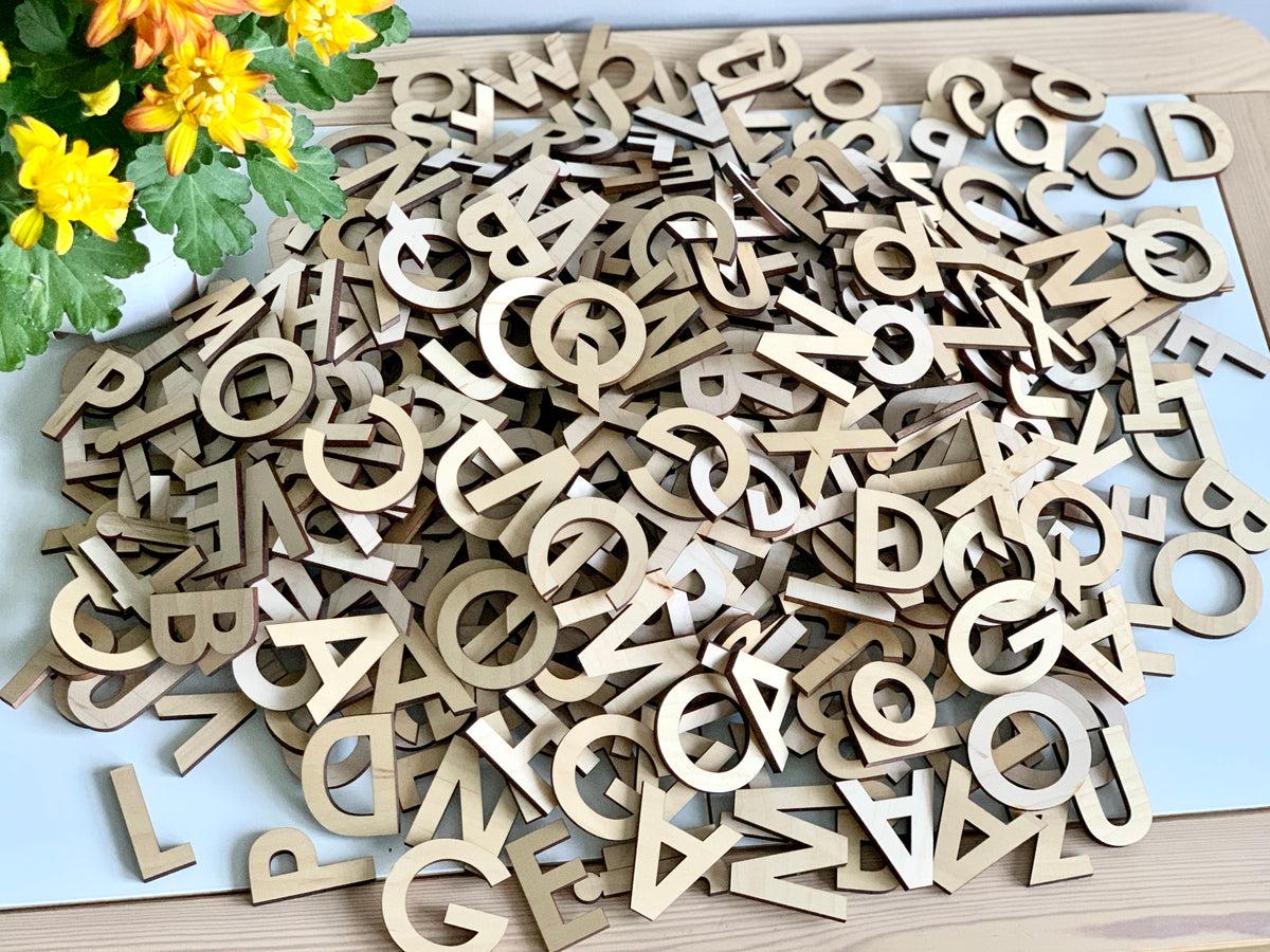 Pile of natural wooden uppercase alphabet letters scattered on white surface with yellow flowers nearby