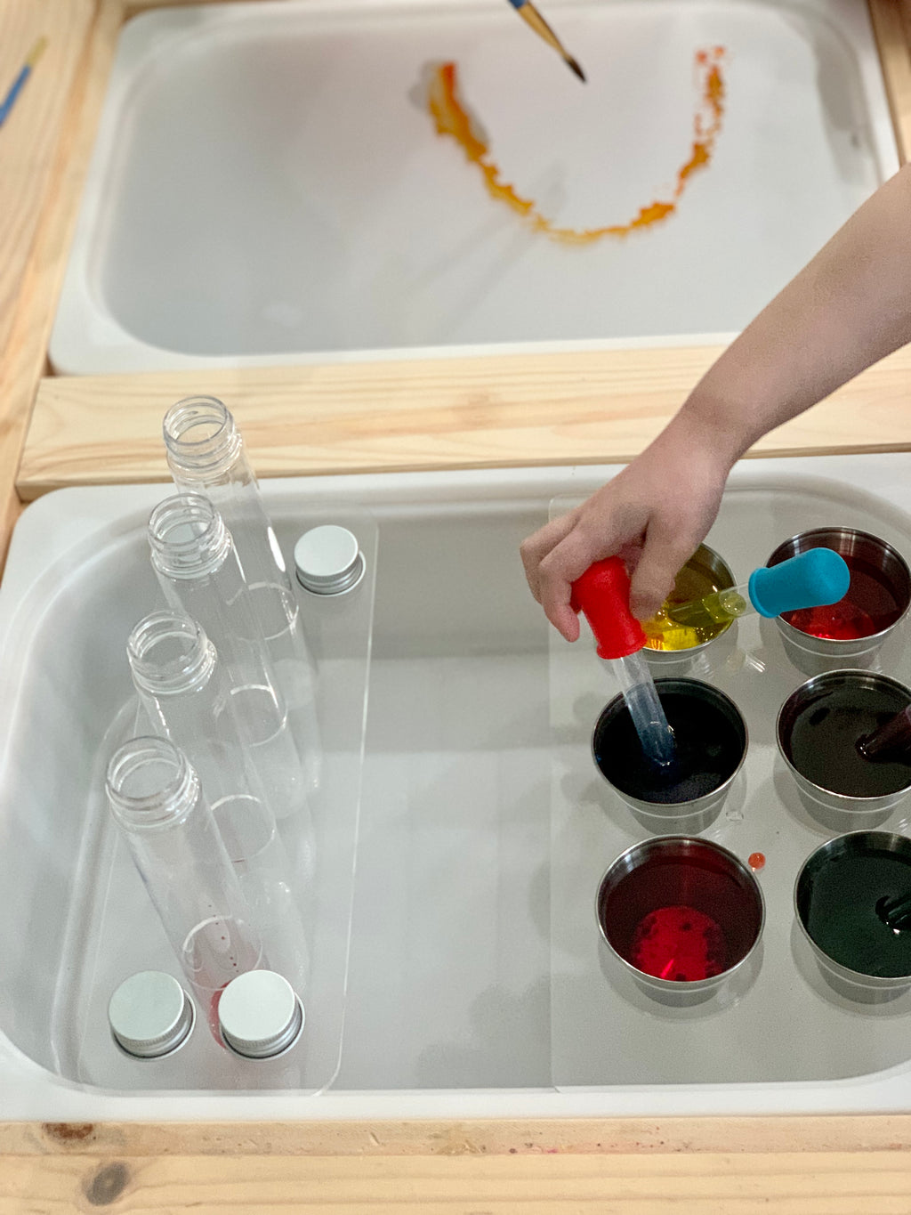 Clear acrylic Flisat table insert with plastic test tubes and metal lids beside metal cups with colored liquids and child’s hand using dropper