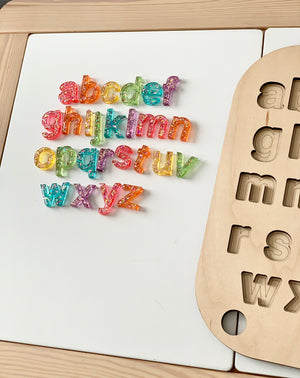 Lowercase resin alphabet letters in multicolor glitter beside a wooden Flisat puzzle board with matching letter cutouts on white table