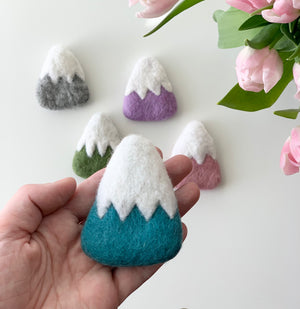 Handmade blue felt mountain with white peak held in hand with purple, green, pink, and gray felt mountains on white background