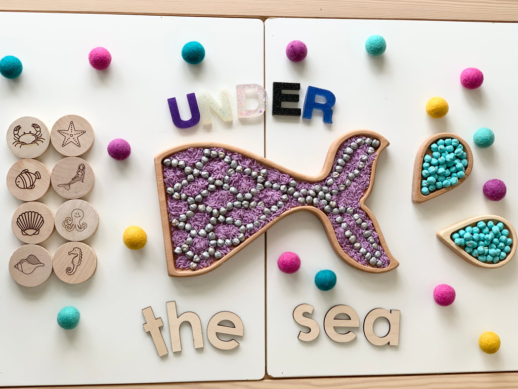 Mermaid tail sensory tray filled with silver and purple beads with wooden sea-themed tokens and colorful felt balls on white surface