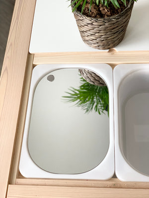 Small mirrored acrylic Flisat table bin lid with rounded corners reflecting green plant and woven basket above wooden frame