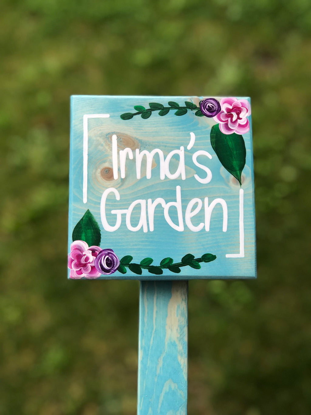 Square wooden garden sign painted light blue with white cursive letters and hand-painted pink and purple flowers on natural wood grain