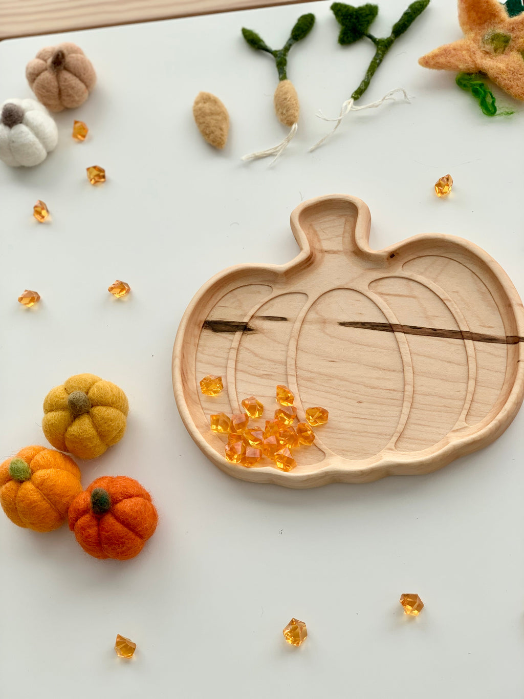 Wooden pumpkin-shaped sensory tray with engraved pumpkin design surrounded by small felt pumpkins and play gems on white surface