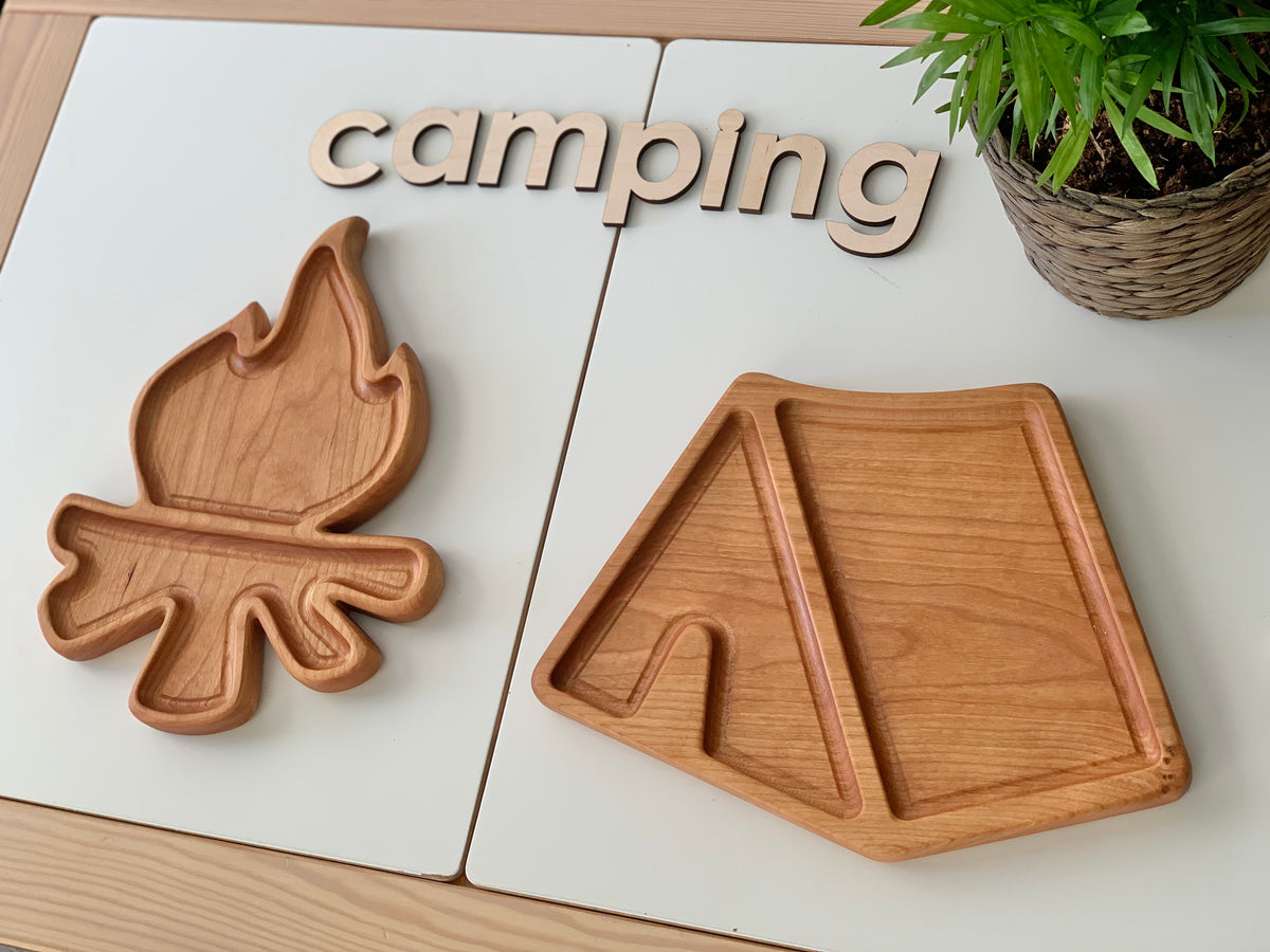 Two wooden sensory trays shaped like a campfire and a tent on white placemats with wooden camping letters