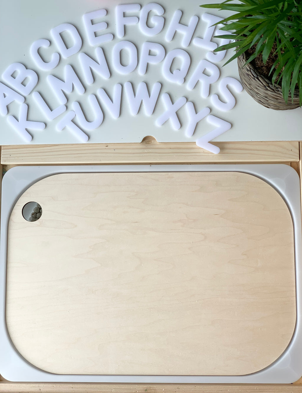 Natural wood Flisat table bin lid insert with rounded edges on light wooden frame with white alphabet letters and green plant nearby