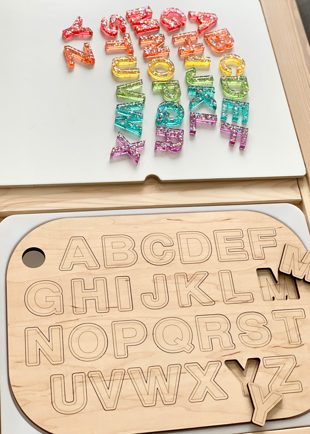 Wooden uppercase alphabet puzzle board with colorful resin lowercase letters scattered on white surface above it